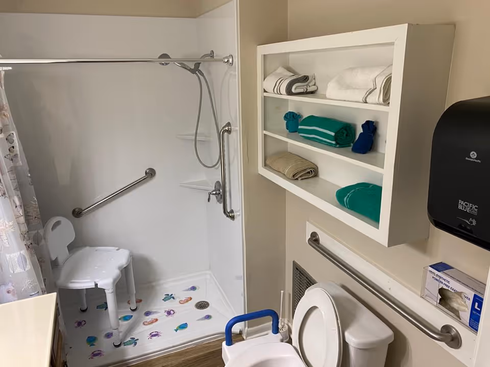 A bathroom with a walk-in shower equipped with a white shower chair and grab bars. The shower floor has colorful sea creature decals. Next to the shower is a toilet with a blue support handle and grab bar on the wall. Above the toilet is a white shelf holding neatly folded towels in white, teal, and beige. A black paper towel dispenser and a box of gloves are mounted on the wall.