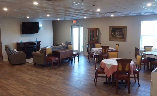 A spacious common area with wooden flooring featuring several round tables covered with tablecloths and surrounded by wooden chairs with cushioned seats. There are two armchairs and a sofa arranged near a wall-mounted flat-screen TV. A glass-front cabinet and a framed painting hang on the walls. Large windows allow natural light to brighten the room.