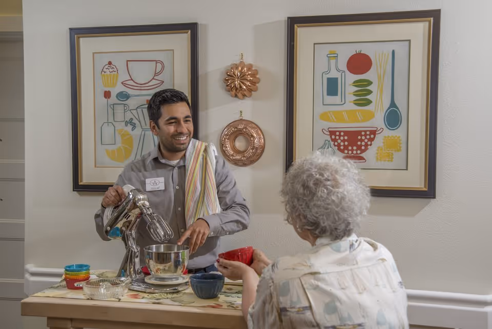 A man with a name tag labeled Joshua is smiling and using a hand mixer in a kitchen setting while an elderly woman with curly gray hair sits across the table holding a red bowl. The wall behind them features two framed pictures of kitchen-themed illustrations and two decorative copper molds.