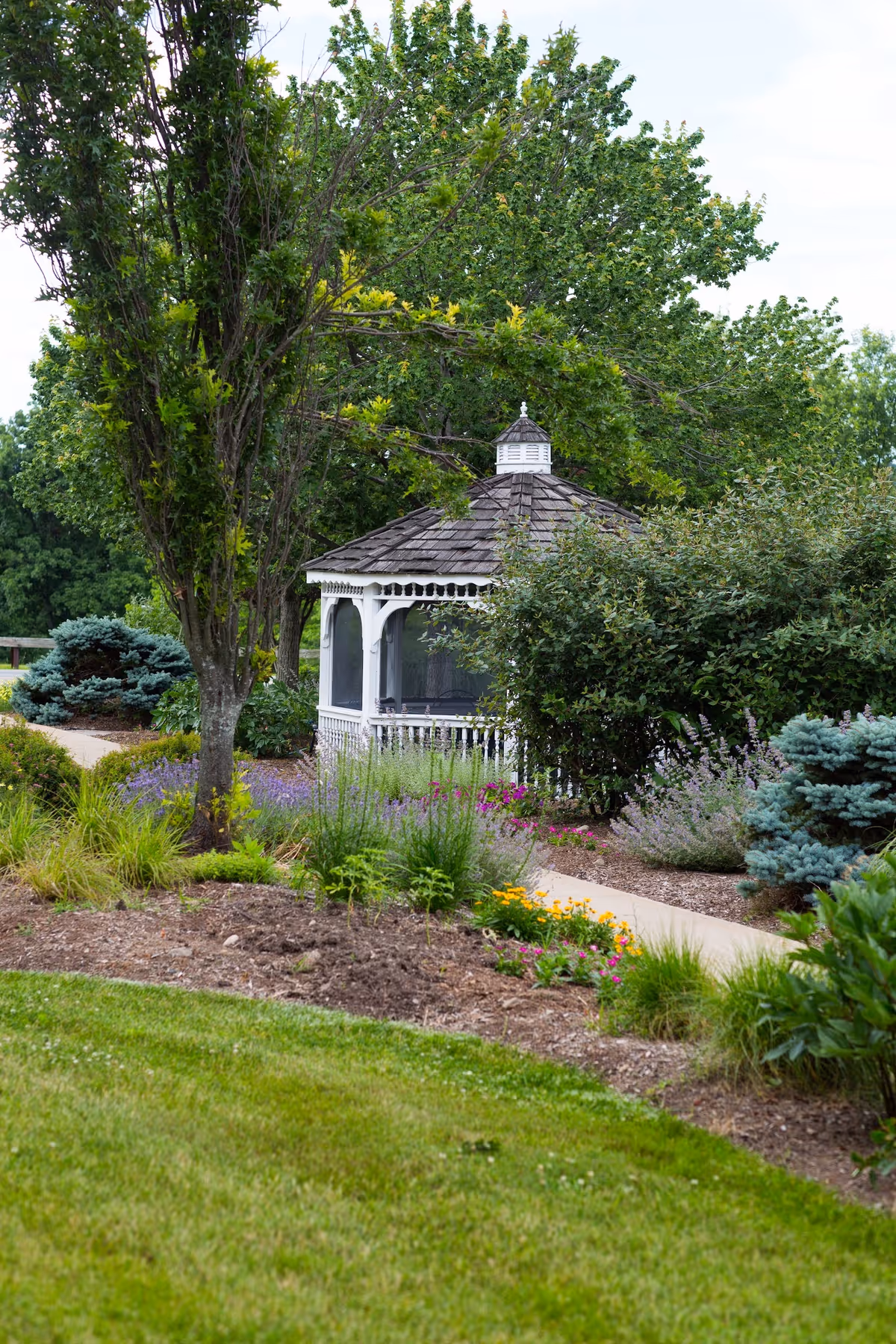 A white wooden gazebo with a shingled roof is surrounded by lush greenery, colorful flowers, and various shrubs in a well-maintained garden area with a paved walkway.