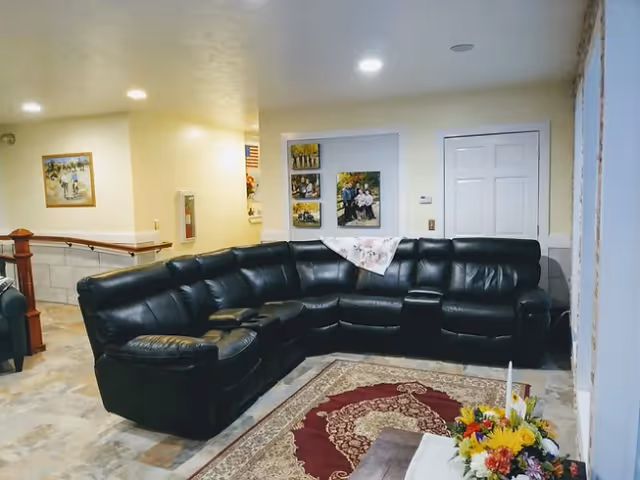 Interior view of a living room area featuring a large black leather sectional sofa arranged around a patterned area rug. The walls are light-colored with framed photos and artwork hanging. There is a floral arrangement on a table in the foreground and recessed ceiling lights providing illumination.