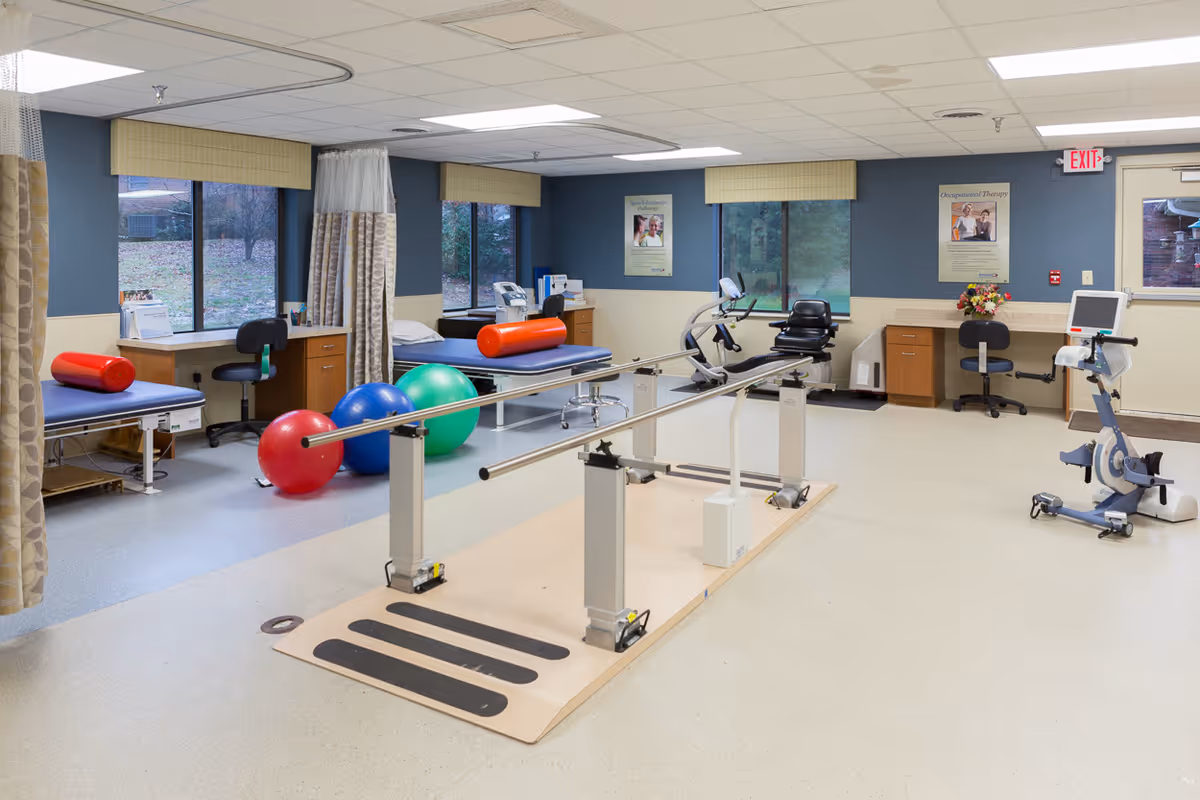 A rehabilitation therapy room with parallel bars for walking exercises in the center, colorful exercise balls on the floor, two treatment tables with red cylindrical bolsters, exercise bikes, desks with chairs, and windows showing an outdoor view.