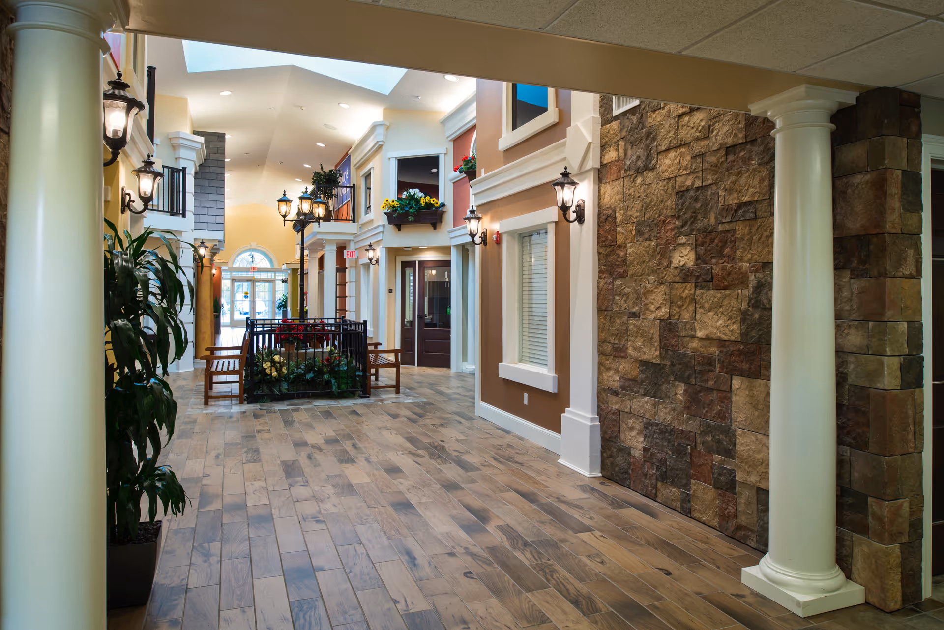 Indoor hallway of a senior living facility designed to look like an outdoor street scene, featuring wooden flooring, white columns, stone accent wall, decorative street lamps, potted plants, benches, and small balcony-style window boxes with flowers.