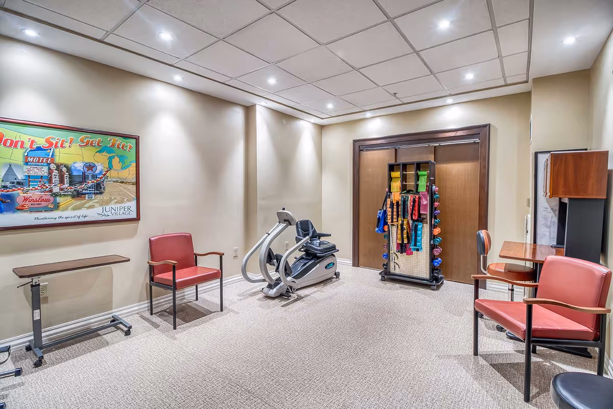 A small exercise room with beige walls and carpeted floor, featuring a stationary recumbent exercise bike, two red chairs, a small adjustable table, and a rack holding various colorful exercise bands and weights. A framed poster on the wall reads 'Don't Sit! Get Fit!' with the Juniper Village logo.