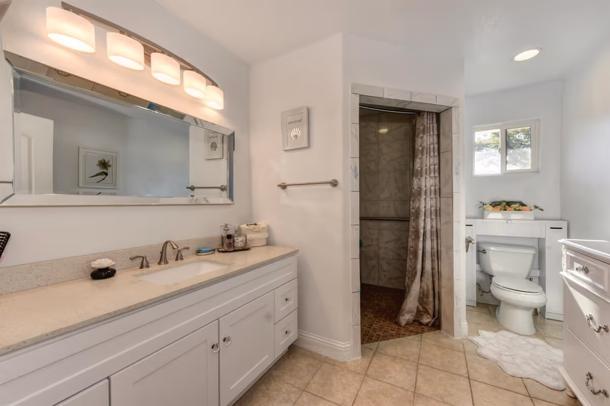 Bright bathroom with a long white vanity and mirror on the left, a tiled walk-in shower with curtain in the center-right, and a toilet under a small window on the far right.