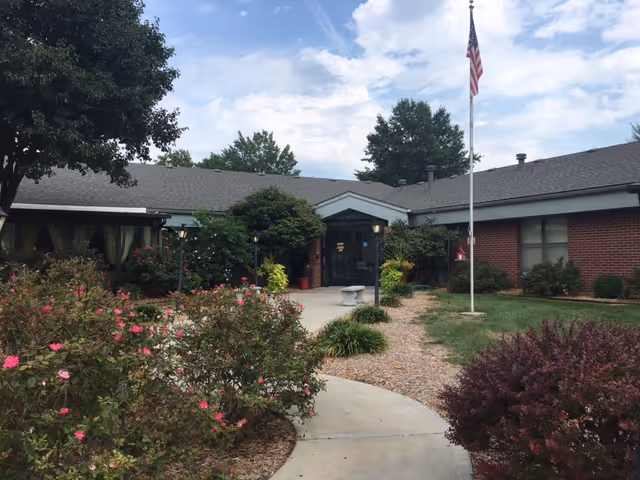 Front entrance of a single-story brick health facility with a curved concrete walkway, landscaped shrubs and an American flag on a flagpole.