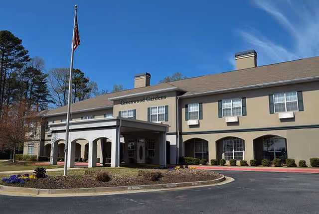 Two-story beige building with a covered main entrance, American flag, landscaped circular driveway, and rows of windows under a blue sky.