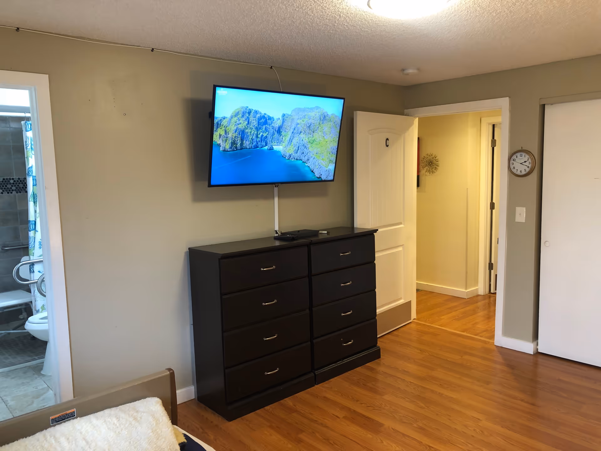 Interior view of a room with wooden flooring, a dark wooden dresser with multiple drawers, and a flat-screen TV mounted on the wall above the dresser displaying a scenic image of a blue sea and green cliffs. To the left, there is a doorway leading to a bathroom with tiled walls and a shower curtain. To the right, there is an open door labeled with the letter 'C' and a hallway with light-colored walls and a clock on the wall.