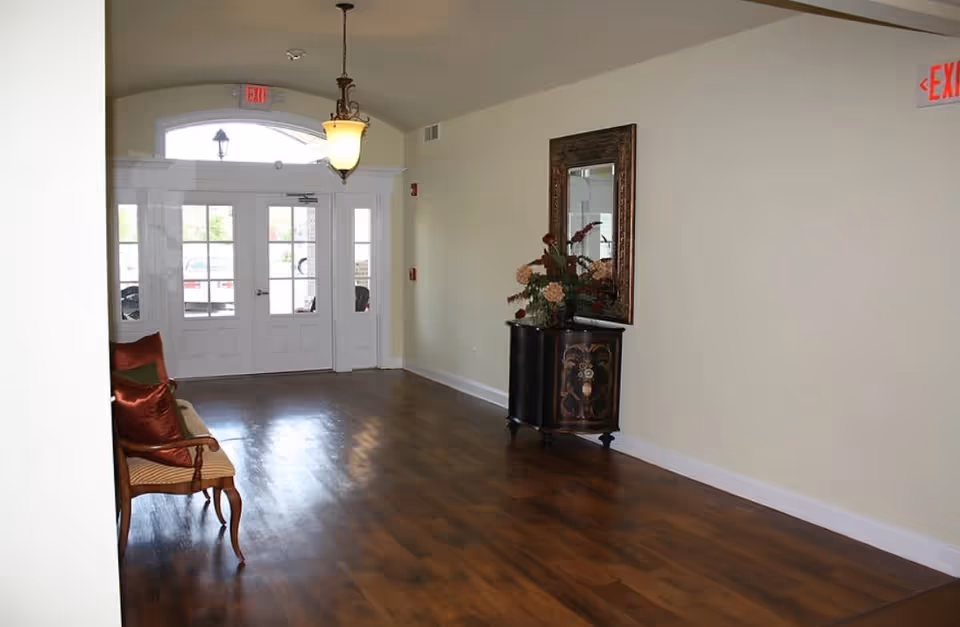 Entrance area of Stonecrest Assisted Living Facility with wooden flooring, a decorative cabinet with a floral arrangement and mirror on the right wall, a bench with cushions on the left, and double glass doors letting in natural light.