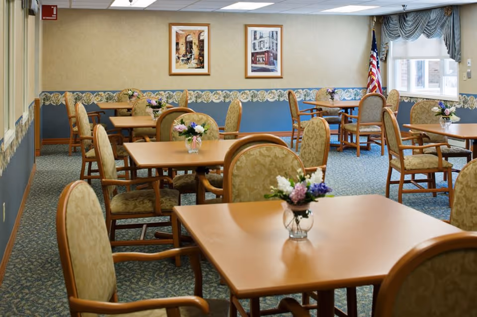 A dining room with multiple square wooden tables, each surrounded by upholstered chairs with floral patterns. Small flower arrangements in glass vases are placed on each table. The room has blue and beige wallpaper with a floral border, carpeted floor, framed artwork on the walls, and an American flag near a window with draped curtains.