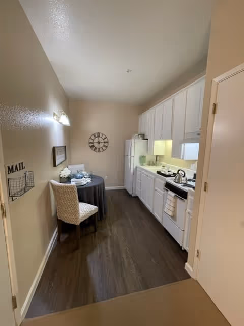 A compact kitchen and dining area with white cabinets, a white refrigerator, and a stove with a kettle on it. A small round table covered with a dark tablecloth is set with plates and napkins, accompanied by a single upholstered chair. The walls are beige, and there is a decorative wall clock and a mail holder on the left wall.
