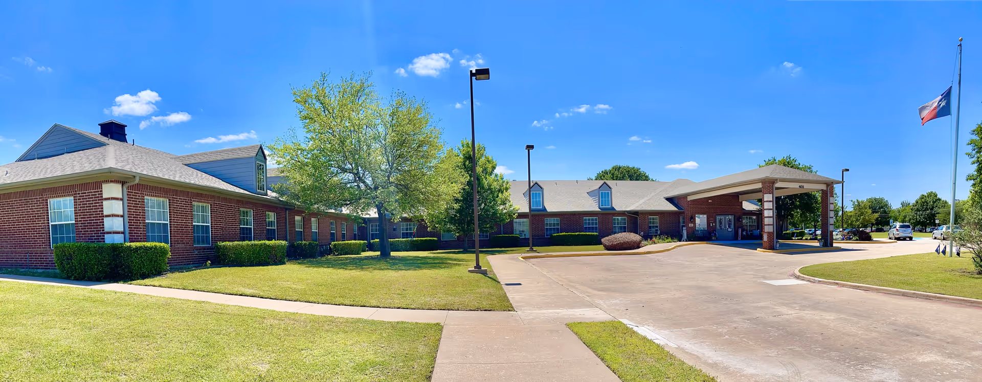 Exterior view of Morada Waxahachie facility showing a single-story brick building with multiple windows, a covered entrance, a driveway, green lawns, trees, and a flagpole with the Texas state flag against a clear blue sky.