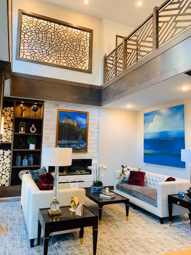A bright and modern senior living facility common area with two white sofas adorned with colorful cushions, a dark wooden coffee table, and side tables with lamps. The room features a fireplace with a framed painting above it, decorative shelving with logs and ornaments, and a large abstract blue painting on the wall. The space has a high ceiling with a mezzanine level and decorative railing.