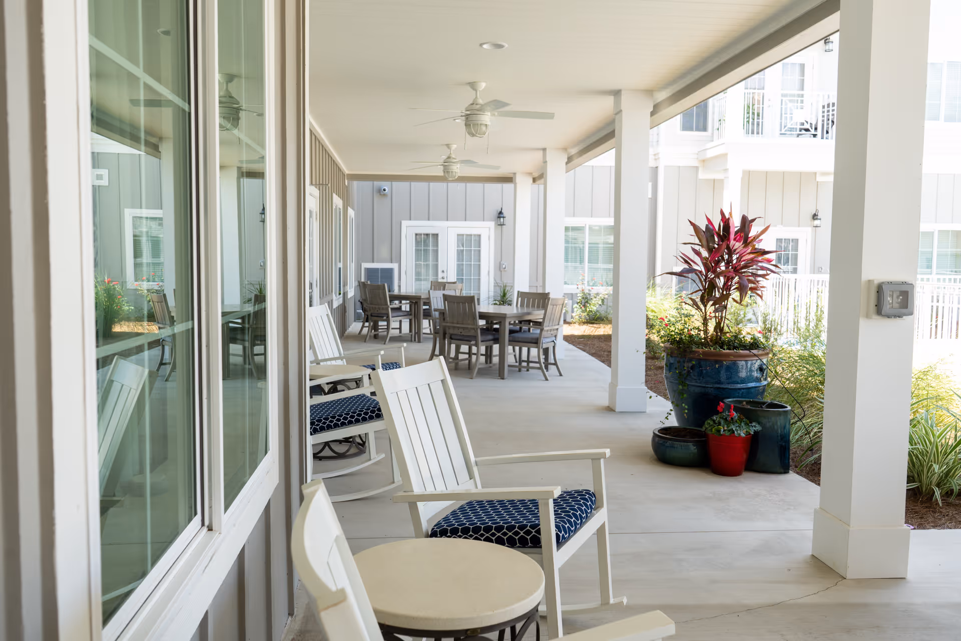 Covered outdoor patio area with white rocking chairs featuring blue patterned cushions, round tables, ceiling fans, and several potted plants. The patio is adjacent to a building with large windows and light gray siding.