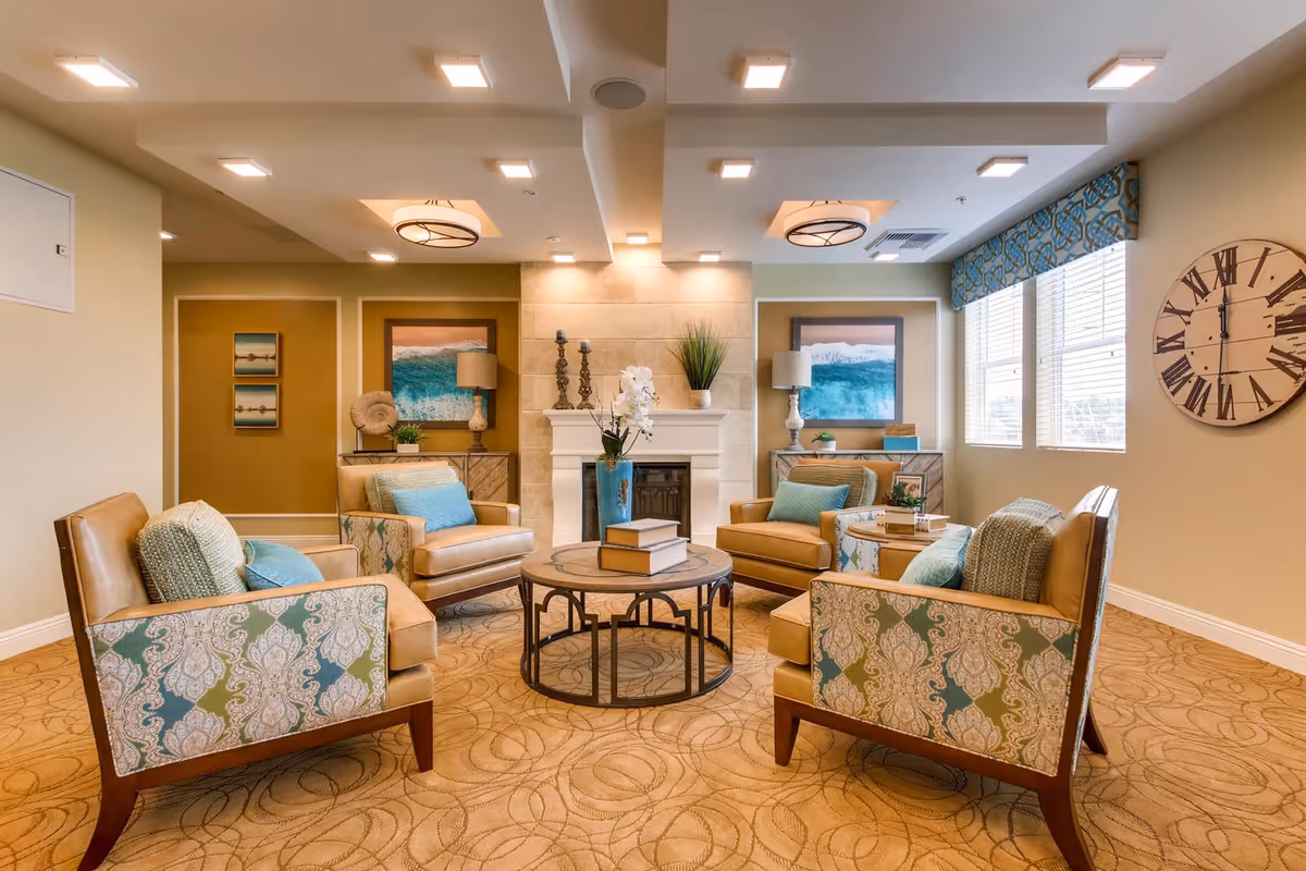 A cozy living room area with four patterned armchairs arranged around a round wooden coffee table. The room features a fireplace with decorative items on the mantel, two side tables with lamps, framed ocean-themed artwork, a large wall clock, and windows with blinds and patterned valances letting in natural light.