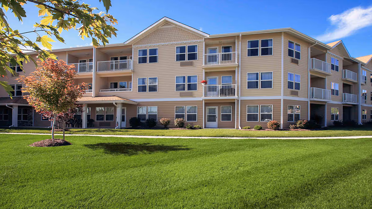 Exterior view of a three-story senior living facility building with beige siding, multiple windows, and balconies. The building is surrounded by a well-maintained green lawn and a few small trees under a clear blue sky.