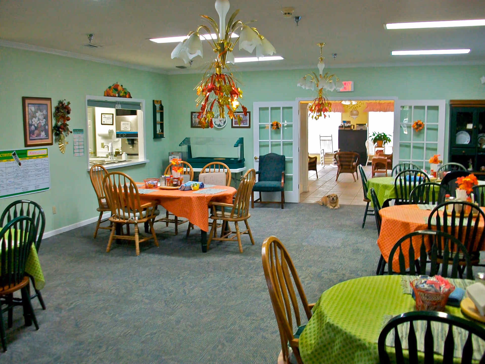 A dining room in a senior living facility with several round tables covered with orange and green tablecloths. Wooden chairs surround the tables, and there are decorative light fixtures hanging from the ceiling. The walls are painted light green and decorated with framed pictures and wall hangings. A small dog is lying on the floor near a doorway leading to another room with additional seating and rocking chairs.