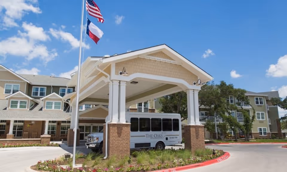 Front entrance of The Oaks senior living building with a covered porte-cochere, flags, and a shuttle van parked underneath.