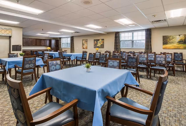 Spacious dining room with square tables draped in blue tablecloths and wooden chairs arranged under ceiling lights.