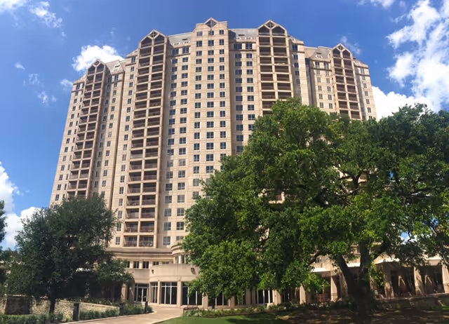 Tall multi-story residential building with numerous balconies, surrounded by large green trees under a blue sky with some clouds.