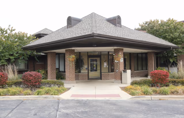 Front exterior view of a single-story brick building with a covered entrance supported by four brick columns. The entrance has glass doors and windows, surrounded by landscaped bushes and plants. The building number 1136 is visible above the entrance.