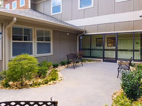 A small paved courtyard with benches, rock-bordered plant beds and a glass doorway along the exterior of a senior living building.