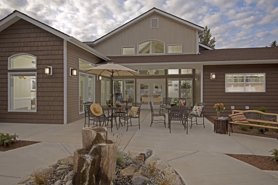 Outdoor patio area of a senior living facility with metal chairs and tables, one table shaded by a large umbrella. A small water fountain with rocks is in the foreground. The building has brown siding with multiple windows and glass doors leading inside. The sky is partly cloudy.