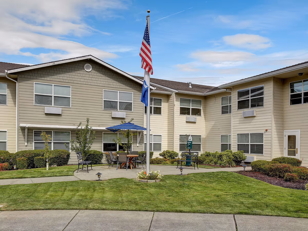 Courtyard and front exterior of a two-story senior living building with a flagpole, patio seating, and landscaping.