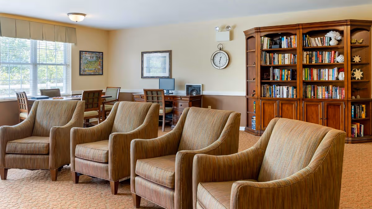 A cozy sitting area with four upholstered armchairs arranged in two rows facing each other. Behind the chairs, there is a wooden bookshelf filled with books and decorative items. A desk with a computer and a chair is positioned against the wall, which is decorated with framed pictures and a round wall clock. Large windows with blinds allow natural light to brighten the room.