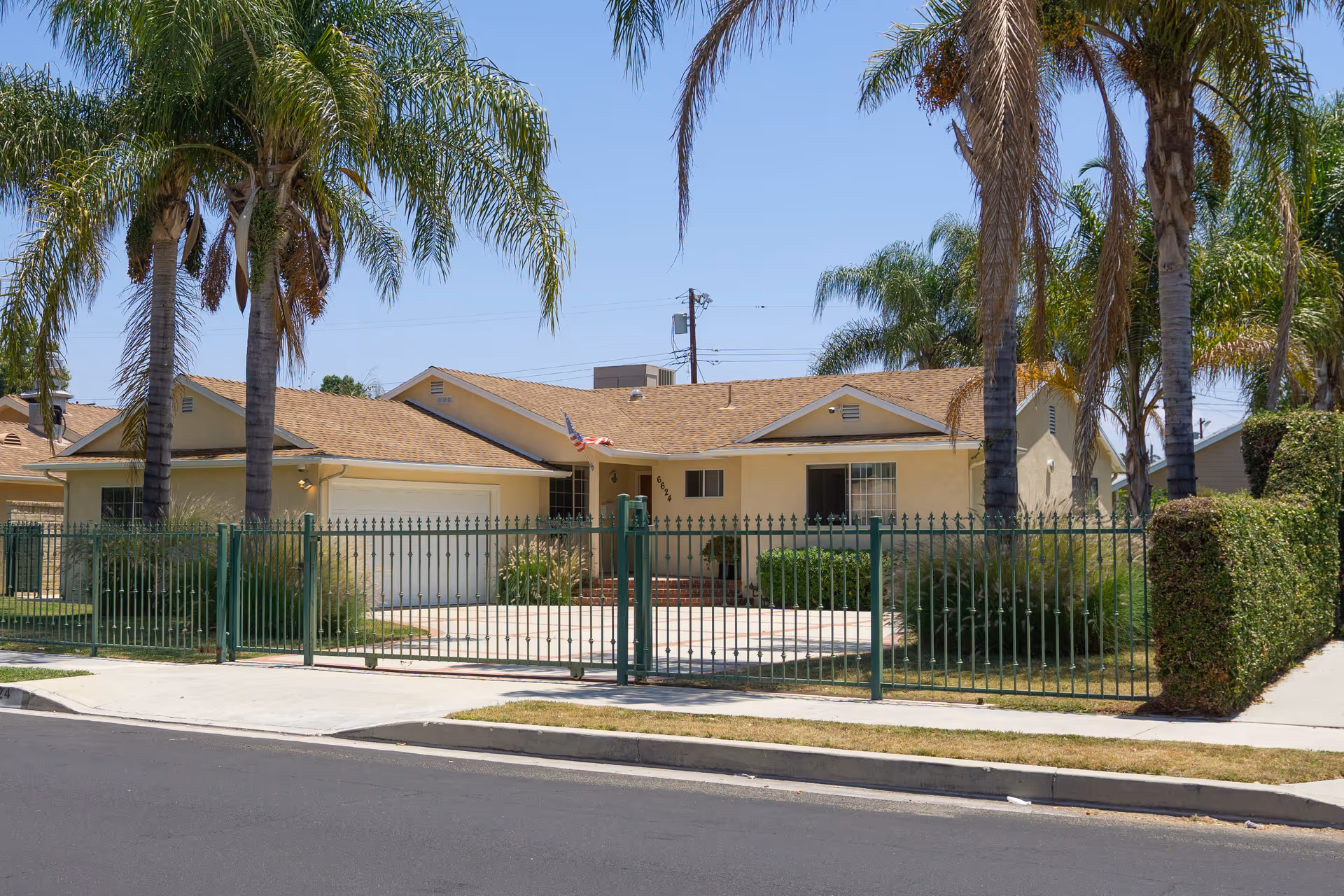 Single-story house with a beige exterior and a brown shingle roof, surrounded by a green metal fence. There are several tall palm trees in the front yard and an American flag mounted near the entrance. The house has a two-car garage and a small front porch with steps leading up to the door. The sky is clear and blue.