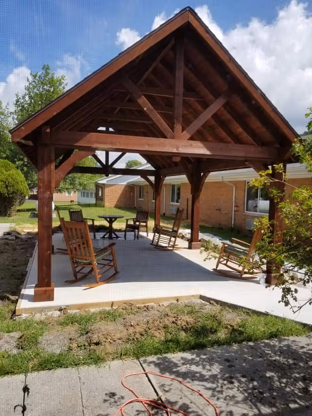 Outdoor wooden gazebo with a pitched roof and four supporting posts, situated on a concrete patio. Under the gazebo are four wooden rocking chairs and a small round table. The area is surrounded by grass, bushes, and a brick building in the background under a partly cloudy sky.