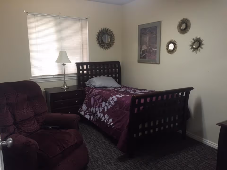 A small bedroom with a single bed covered in a purple floral comforter, a gray pillow, a wooden nightstand with a lamp, a maroon recliner chair, a window with closed blinds, and decorative mirrors and a framed picture on the wall.