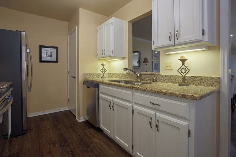 A modern kitchen with white cabinets, granite countertops, a stainless steel refrigerator, dishwasher, and stove. The kitchen has under-cabinet lighting and a decorative candle holder on the counter. The floor is dark wood, and there is a framed picture on the wall near a closed door.