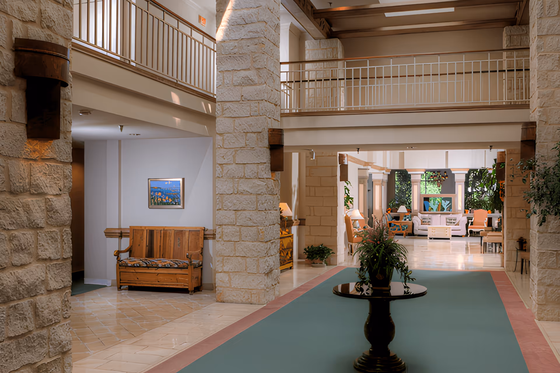 Spacious stone-columned lobby with a central plant-topped table, seating areas in the background, and an upper walkway.