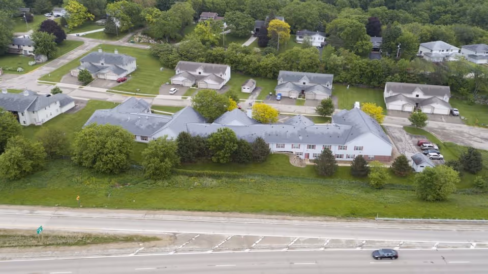 Aerial view of McFarland Villa Assisted Living facility showing a large building with a gray roof surrounded by trees and greenery. The facility is adjacent to a highway with a few cars visible. Residential houses and additional buildings are seen behind the facility.