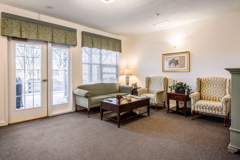 A cozy living room area with a green upholstered sofa, two patterned armchairs, a wooden coffee table with books and a small plant, a side table with a lamp and a plant, and a framed picture on the wall. There are large windows and a glass door with green valances, letting in natural light.