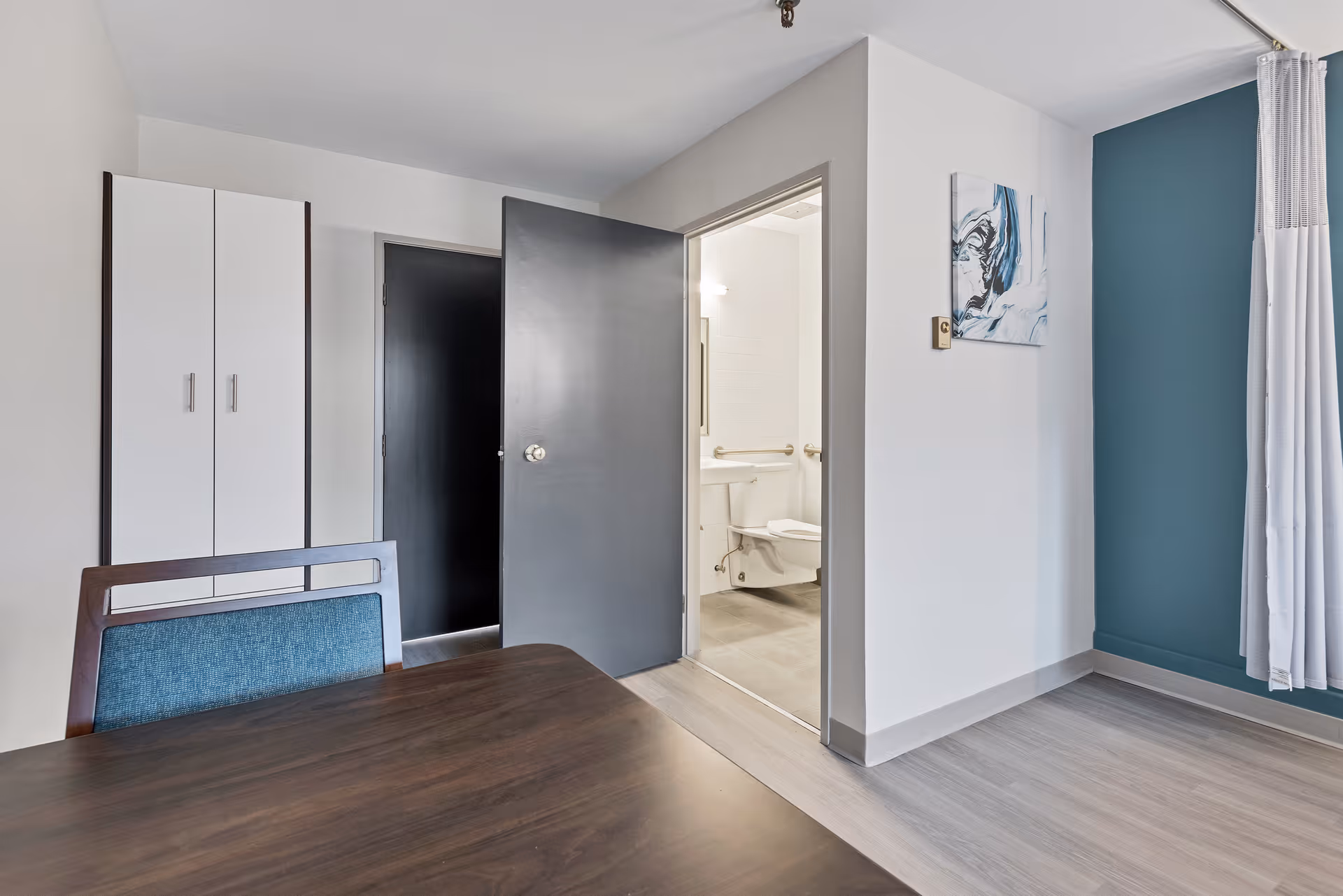Interior view of a room with a wooden table and a chair with blue upholstery in the foreground. There is a white cabinet against the wall and an open door leading to a bathroom with a toilet and grab bars. The walls are painted white and teal, and there is a piece of abstract artwork on the wall.