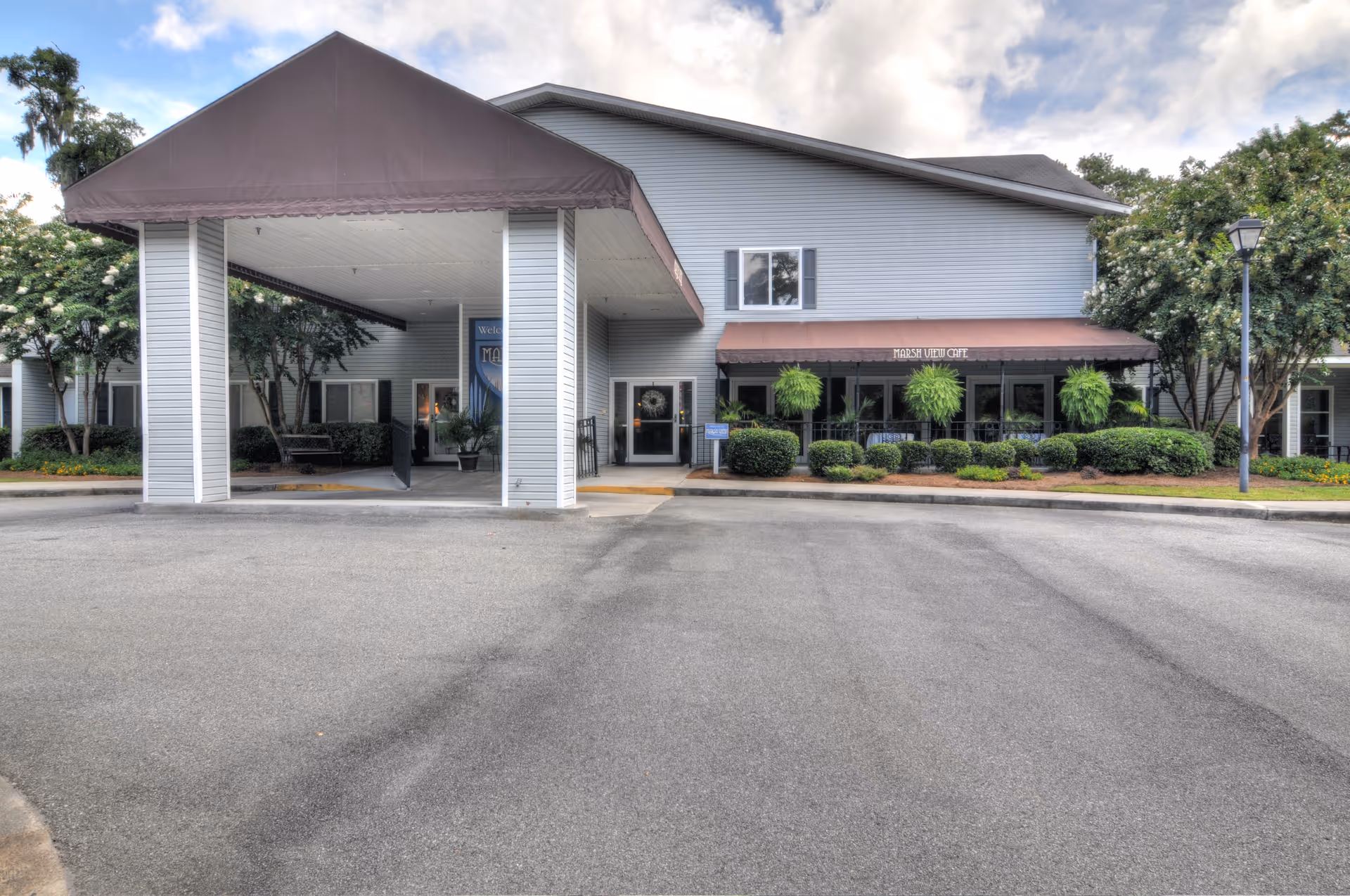 Front entrance and porte-cochère of a two-story senior living building with awnings, shrubs, and a paved driveway.