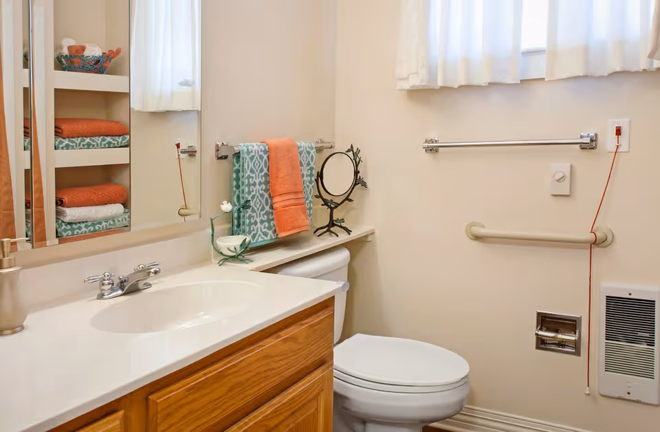 A clean bathroom with a white sink and wooden cabinet, a toilet, a small shelf above the toilet holding a decorative mirror and a small flower pot. There are colorful towels in orange and teal hanging on a towel rack and neatly folded on shelves beside a mirror. The walls are light beige, and there is a window with white curtains. Safety grab bars are installed near the toilet and a wall-mounted heater is visible.