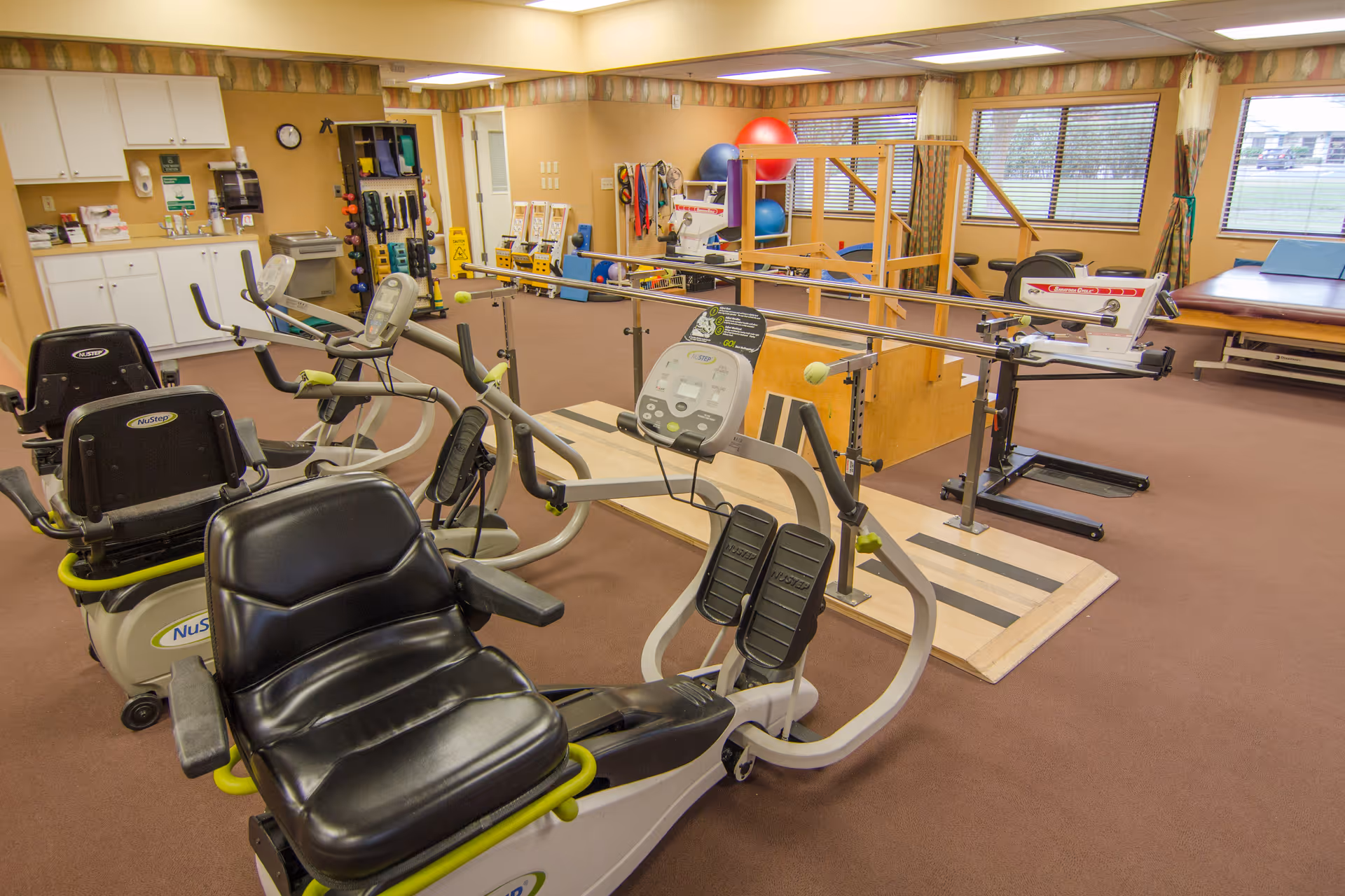 Interior view of a physical therapy or rehabilitation room with exercise equipment including recumbent bikes, parallel bars for walking practice, therapy balls, and other rehabilitation tools. The room has beige walls, carpeted floor, large windows with blinds, and cabinets along one wall.