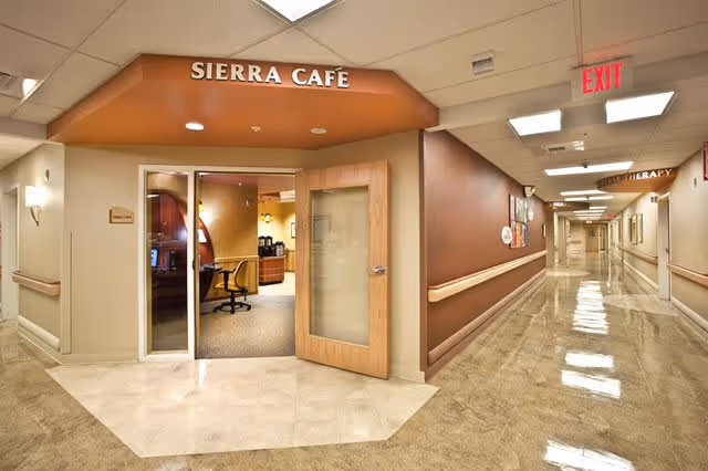 Interior view of a hallway in a senior living facility with a sign above a doorway reading 'SIERRA CAFE'. The hallway has polished floors, beige walls with wooden handrails, and ceiling lights. The cafe entrance has glass doors and a wooden door that is open, revealing a seating area with chairs and tables inside. There is an exit sign visible at the end of the hallway.