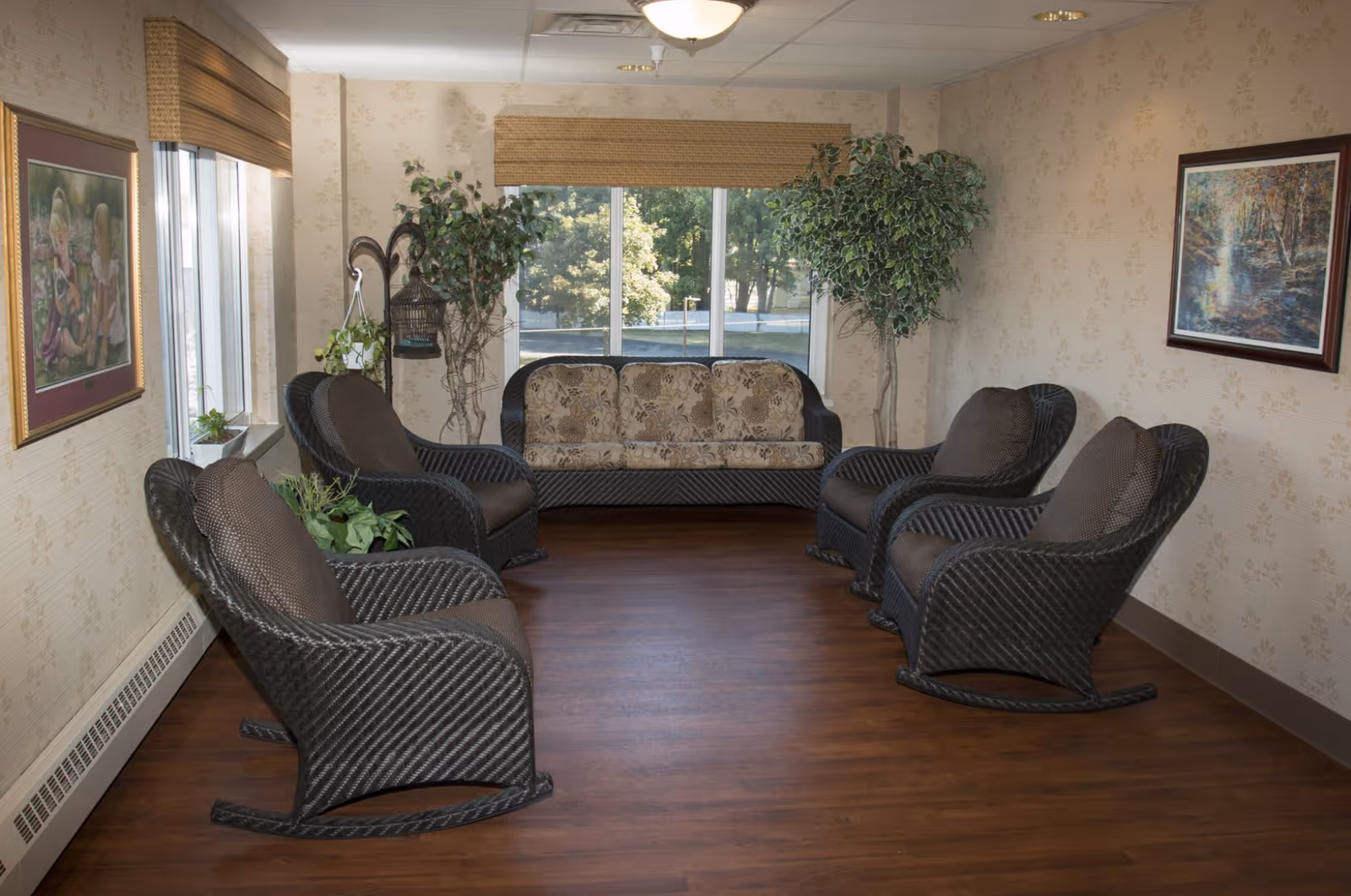 A cozy sitting area in an assisted living facility with four dark wicker rocking chairs and a matching wicker sofa with floral cushions arranged around a wooden floor. The room has beige wallpaper with a subtle floral pattern, two framed paintings on the walls, two large potted plants, and a large window with a view of trees outside. A ceiling light fixture illuminates the space.
