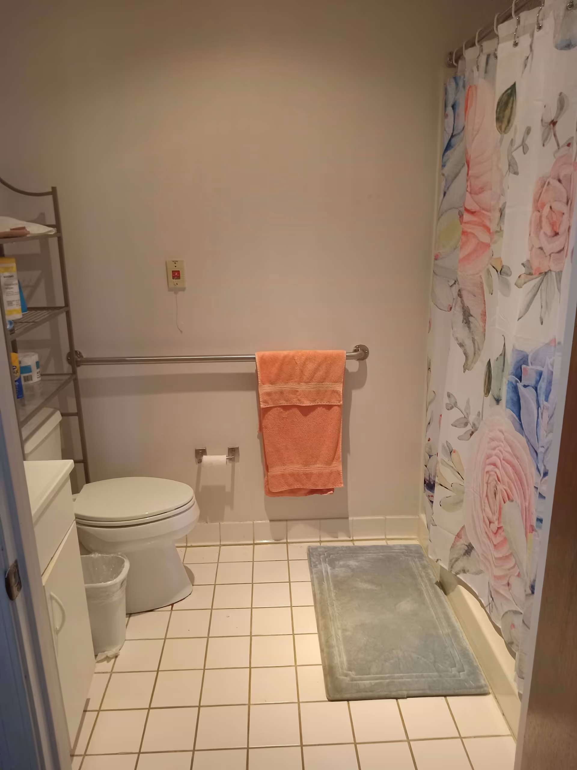 Small bathroom with a toilet, orange towel hanging on a grab bar, floral shower curtain, and a bath mat on a tiled floor.
