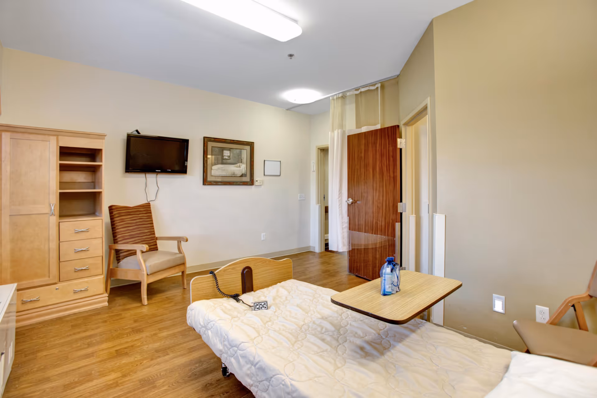 A furnished patient bedroom with a hospital bed and overbed table, wooden wardrobe and chair, wall-mounted TV, and an open doorway.