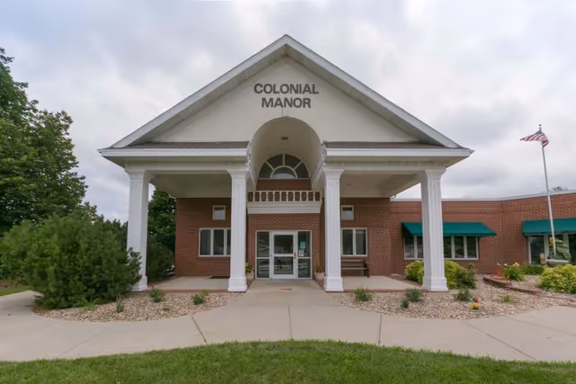 Front exterior view of Colonial Manor building with a covered entrance supported by four white columns, brick walls, green awnings over windows, and an American flag on a flagpole to the right.