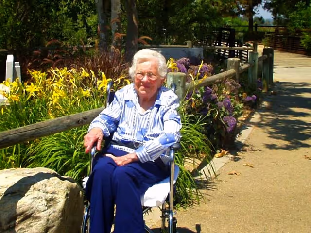 An elderly woman with white hair and glasses sitting in a wheelchair outdoors on a sunny day. She is wearing a blue and white striped shirt and dark blue pants. Behind her is a wooden fence with yellow and purple flowers and green foliage. A paved pathway runs alongside the fence.