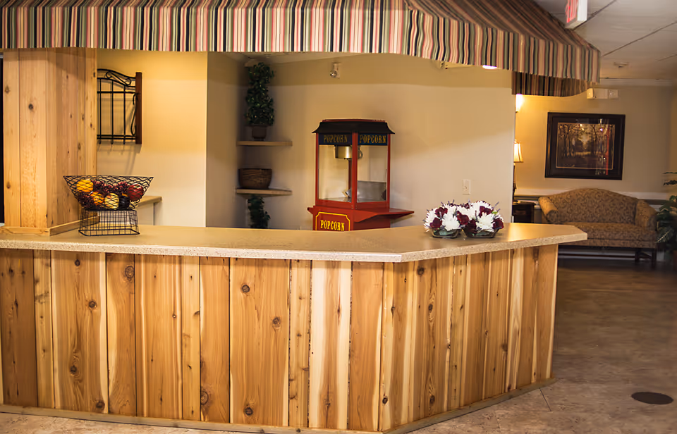 Interior view of a reception or common area with a wooden counter topped with a basket of fruit and a flower arrangement. Behind the counter is a red popcorn machine and corner shelves with decorative plants. In the background, there is a patterned sofa, a lamp, and a framed picture on the wall.