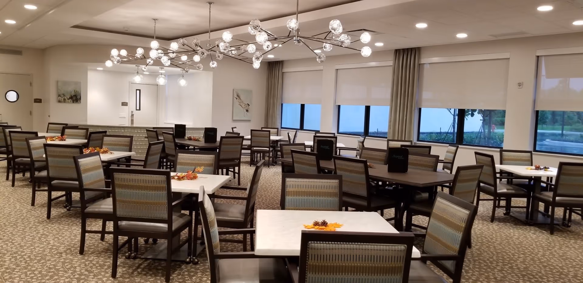 A spacious dining room with multiple tables and chairs arranged neatly. The tables have small autumn-themed centerpieces with leaves and pinecones. Large windows with roller shades allow natural light to enter the room. Modern chandeliers with multiple light bulbs hang from the ceiling, illuminating the space. The carpet has a patterned design, and the walls are decorated with framed artwork.