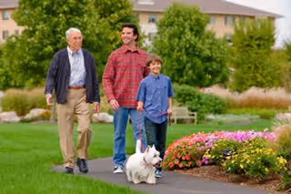 Three people walking a small white dog along a landscaped path in front of a senior living building.