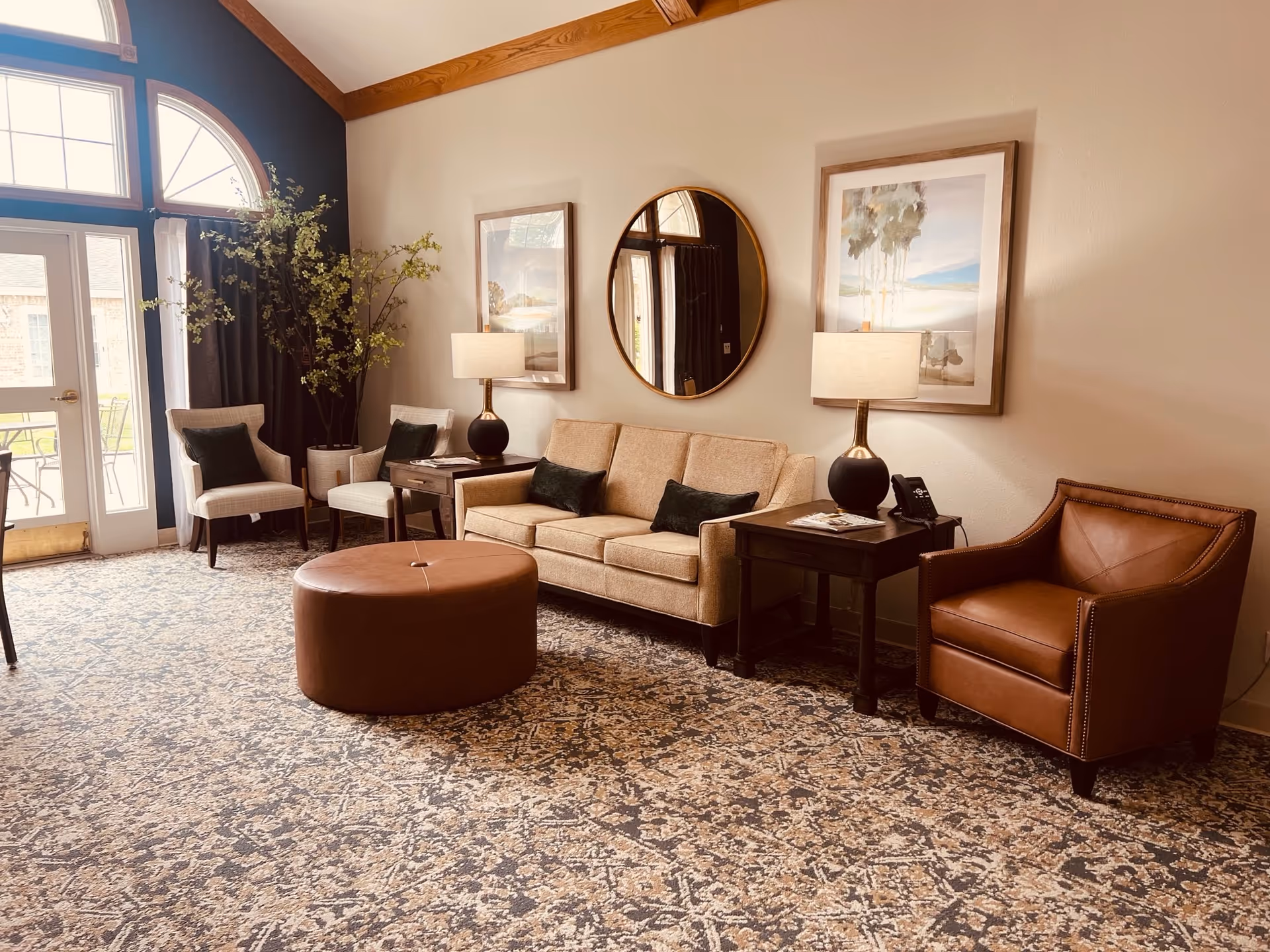 A well-lit living room with a beige sofa, leather armchair, two side chairs, round ottoman, side tables, lamps, wall art and large arched windows.